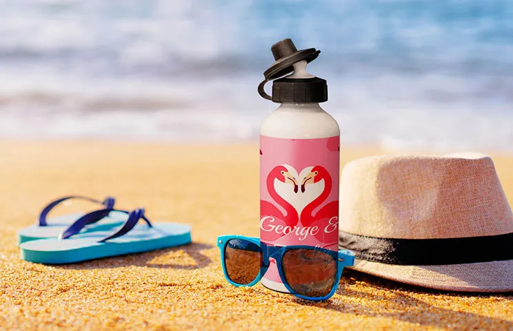 Gepersonaliseerde flamingo waterfles op zandstrand, naast zonnebril & hoed. Perfect zomergeschenk!